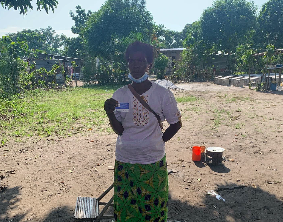 Foto de Micheline em um campo aberto mostrando o cartão por meio do qual recebe recursos do projeto de proteção social para famílias vulneráveis do país.