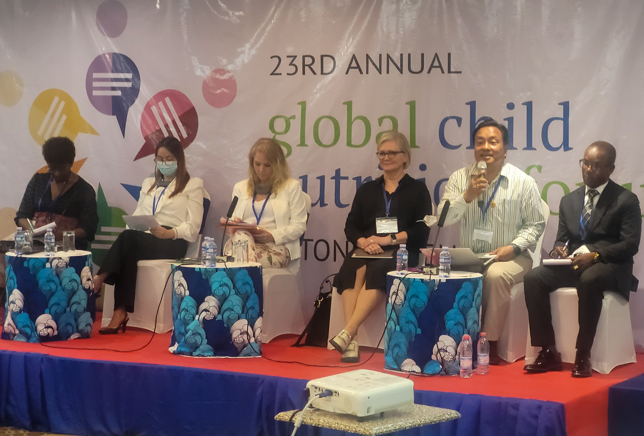 Imagem mostra seis representantes sentados em um palco azul com vermelho com um painel escrito "23rd annual global child nutrition fund" no fundo