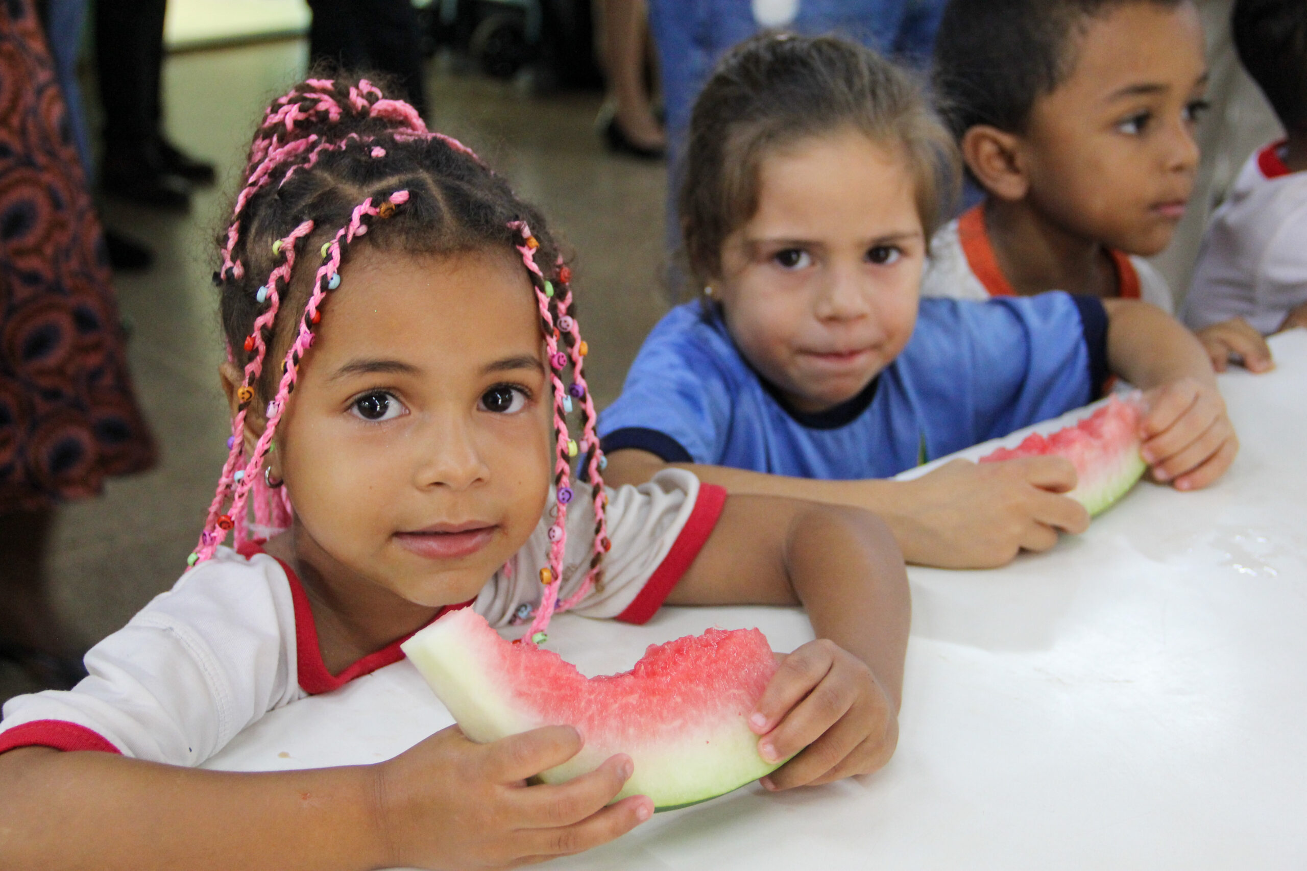 The photo shows three children, a black girl with pink braids in her hair eating a watermelon wearing a white blouse with red details from the school uniform, a white girl with straight hair eating a watermelon wearing a blue blouse, and a black boy wearing a white blouse with orange details from the school uniform.