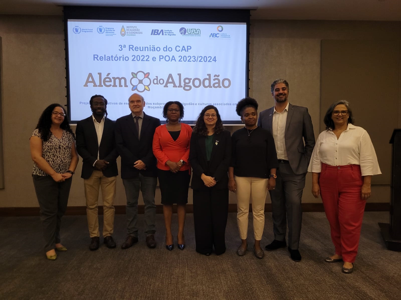 Grupo de pessoas, homens e mulheres, usando roupas formais em uma sala de reuniões frente a uma projeção que tem a logo do projeto Além do Algodão 