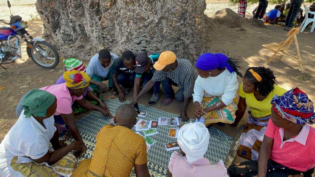 A group of black people wearing colourful clothes squatted in a circle with pictures of food in the centre pointing to some of them.