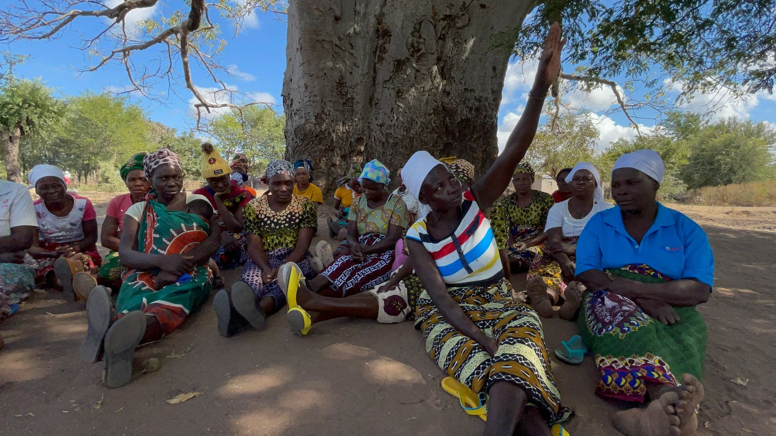 Group of black people wearing colourful clothes sitting around a tree.