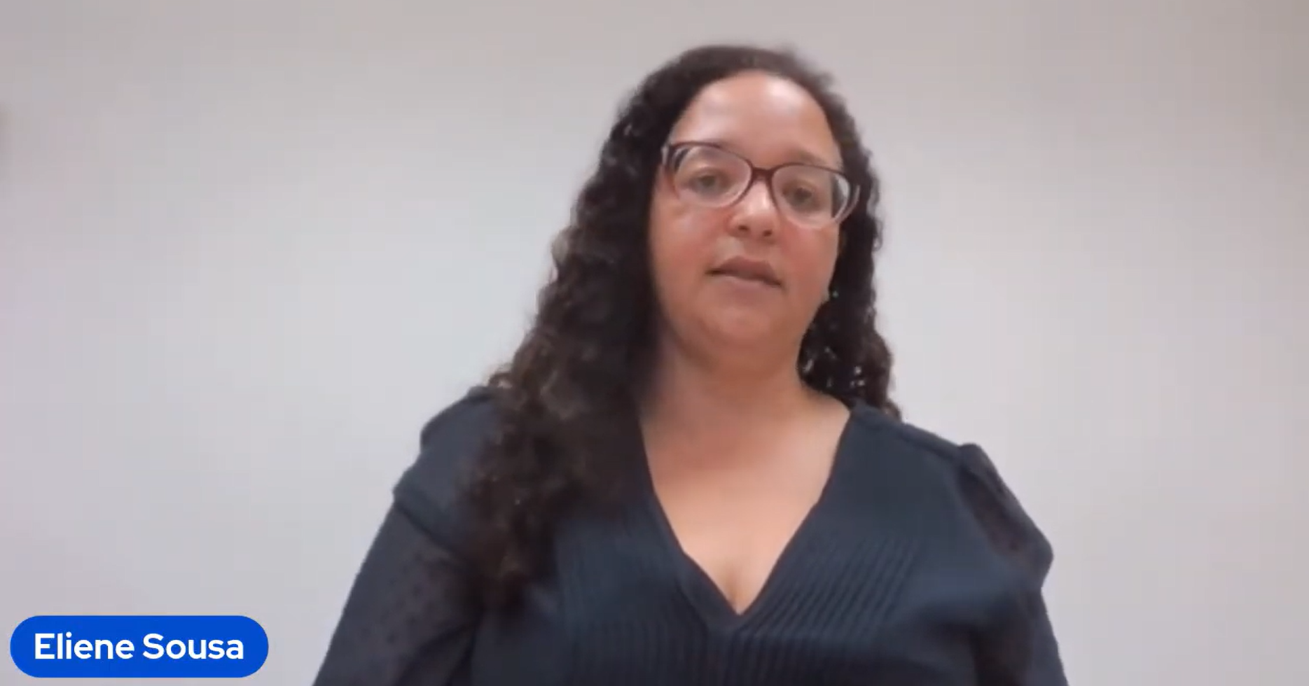 Eliene Souza, a light-skinned black woman with long curly hair wearing a dark blue blouse and glasses in front of a white background.