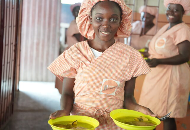 Menina negra sorrindo usando uma touca e um vestido listrados de laranja com branco segura dois pratos de comida.