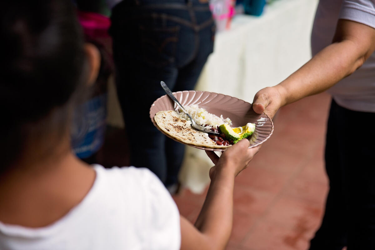 Criança de costas recebendo um prato de comida com uma tortilha, arroz, feijão e abacate de uma pessoa adulta.