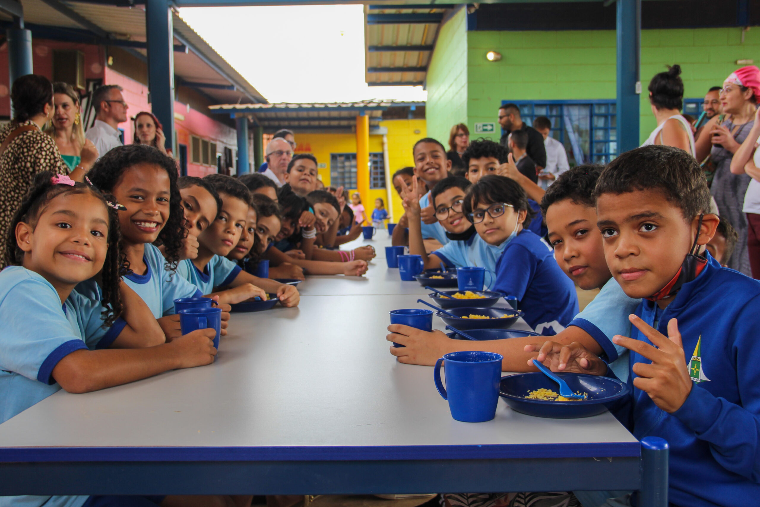 Crianças de uniforme azul comem sua refeição escolar, composta por suco e cuscuz, em uma escola pública brasileira.