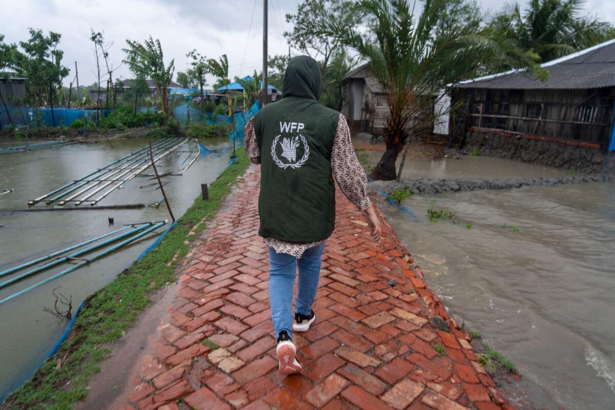 The photo shows a WFP humanitarian worker wearing a dark green waistcoat with the WFP logo in the middle of a flooded area with tents and destroyed buildings around her.