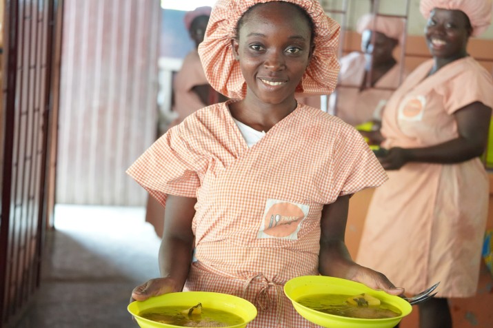 Menina negra sorrindo usando uma touca e um vestido listrados de laranja com branco segura dois pratos de comida.